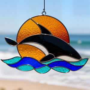 May include: Stained glass suncatcher featuring a black and white orca leaping over blue and teal waves, with an orange sun in the background. The suncatcher is suspended by a silver chain, perfect for a window or wall.