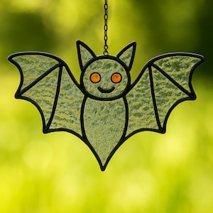 Puede incluir: Decoración de murciélago de vidriera con contorno negro y cadena para colgar. El murciélago tiene una cara sonriente con ojos naranjas y alas texturizadas de color verde claro. El fondo es verde borroso.