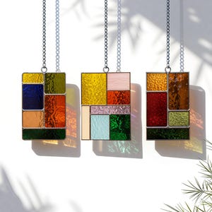 Pode incluir: Três painéis retangulares de vitrais pendurados por correntes prateadas. Cada painel apresenta um design geométrico com várias cores, incluindo azul, laranja, amarelo, verde e vermelho. Os painéis são iluminados, projetando sombras coloridas.