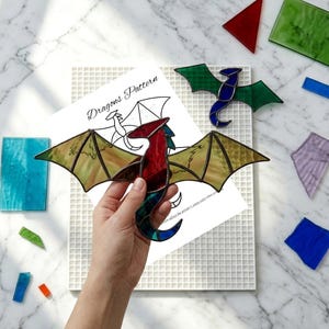 Op de afbeelding: Een glas-in-lood drakenornament met rode, blauwe en getextureerde geelgroene vleugels, gehouden boven een papieren patroon met het label Dragons Pattern. Een kleinere groen-blauwe glas-in-lood draak ligt op een wit rasterbord met glasscherven.