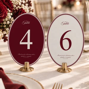 May include: Two oval table number signs, one deep red with a white number 4 and one white with a deep red number 6. Both feature the text Table, Thank you for celebrating with us, MADDIE & CHARLES, and 12.28.2024, supported by gold metal stands.