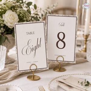 Puede incluir: Dos tarjetas de mesa rectangulares con texto marrón oscuro y un borde fino con un patrón floral de líneas. Una tarjeta dice TABLE Eight, la otra TABLE 8, ambas incluyen los nombres MADDIE & CHARLES y la fecha 12.20.2024.