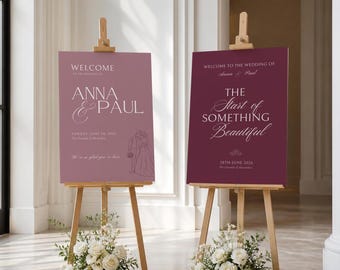 May include: Two rectangular wedding welcome signs on wooden easels. The left sign is mauve with white text reading "WELCOME TO THE WEDDING OF ANNA & PAUL" and a line drawing of a couple. The right sign is dark burgundy with white text reading "THE START OF SOMETHING BEAUTIFUL".