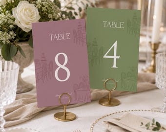 May include: Two rectangular table number cards in mauve and sage green, each featuring the word TABLE, a large numeral, and line art illustrations of chairs and tables. The cards are held upright by small gold metal circular stands.