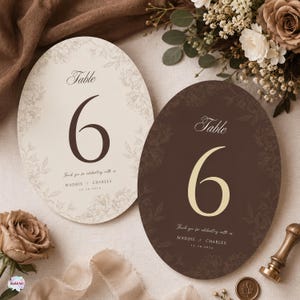 May include: Two oval wedding table number cards featuring the number 6. One card is cream with dark brown text, the other is dark brown with cream text. Both display the text Table, Thank you for celebrating with us, MADDIE & CHARLES, 12.28.2024.