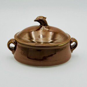 May include: A brown ceramic casserole dish with a lid featuring a bird-shaped knob. The dish has two handles and a glossy finish.