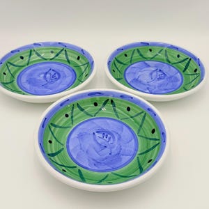 May include: Three ceramic bowls with a white rim, featuring a blue centre with a floral design. The bowls have a green and blue decorative border. The bowls are arranged on a white surface.