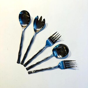 Könnte beinhalten: Set aus vier Servierutensilien aus Edelstahl mit Bambus-Optik-Griffen. Enthält zwei Löffel und zwei Gabeln. Die Utensilien haben eine glänzende Oberfläche und sind auf weißem Hintergrund angeordnet.