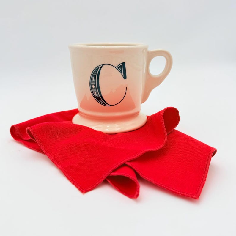 Puede incluir: Una taza color crema con una gran letra "C" azul adornada est&aacute; centrada en un pa&ntilde;o rojo brillante. La taza tiene un asa y una base. El pa&ntilde;o rojo est&aacute; doblado y colocado sobre una superficie blanca.