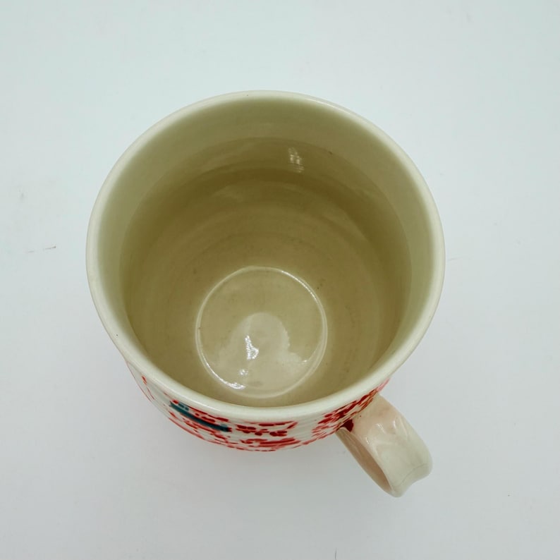 Może przedstawiać: Ceramiczny kubek z kremowym wnętrzem i białym uchwytem. Zewnętrzna strona zdobiona jest wzorem czerwonych kwiatowych wzor&oacute;w z niebieskimi i zielonymi akcentami. Kubek jest widoczny z g&oacute;ry, ukazując jego wnętrze.