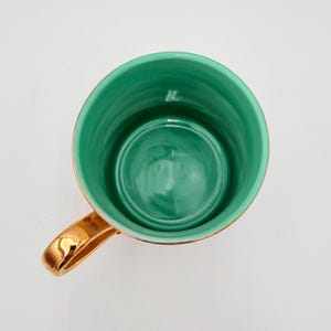 K&ouml;nnte beinhalten: Draufsicht auf eine Keramiktasse mit goldenem Henkel und Rand. Das Innere der Tasse ist gl&auml;nzend t&uuml;rkis. Der Henkel und der Rand sind goldfarben gl&auml;nzend. Die Tasse ist leer und steht auf einer wei&szlig;en Oberfl&auml;che.