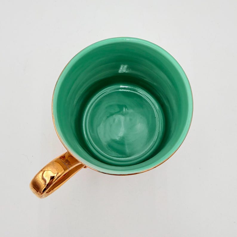 K&ouml;nnte beinhalten: Draufsicht auf eine Keramiktasse mit goldenem Henkel und Rand. Das Innere der Tasse ist gl&auml;nzend t&uuml;rkis. Der Henkel und der Rand sind goldfarben gl&auml;nzend. Die Tasse ist leer und steht auf einer wei&szlig;en Oberfl&auml;che.