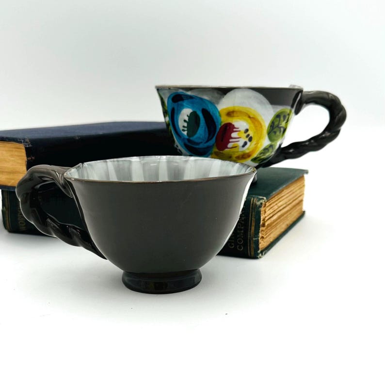 Puede incluir: Dos tazas de t&eacute; decorativas. Una es marr&oacute;n oscuro con un asa texturizada y un interior blanco y gris. La otra es negra con un dise&ntilde;o floral en azul, amarillo y blanco. Ambas tazas descansan sobre libros antiguos.