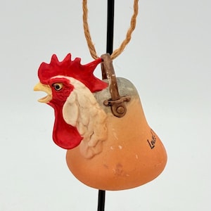 May include: A ceramic rooster ornament with a red comb and wattle, white face, and orange body. The ornament is suspended by a natural fiber cord. The word "Love" is written on the body of the ornament.