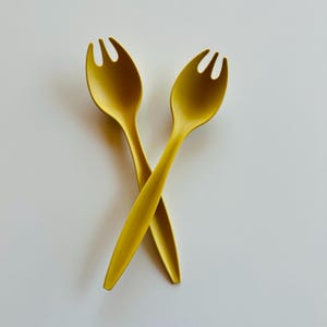 Puede incluir: Dos cucharas de servir de color amarillo dorado con un diseño en forma de tenedor en la cabeza de la cuchara se cruzan sobre un fondo blanco. Las cucharas tienen un acabado liso y mate y un mango largo y delgado. Los utensilios son probablemente para servir ensaladas u otros platos.