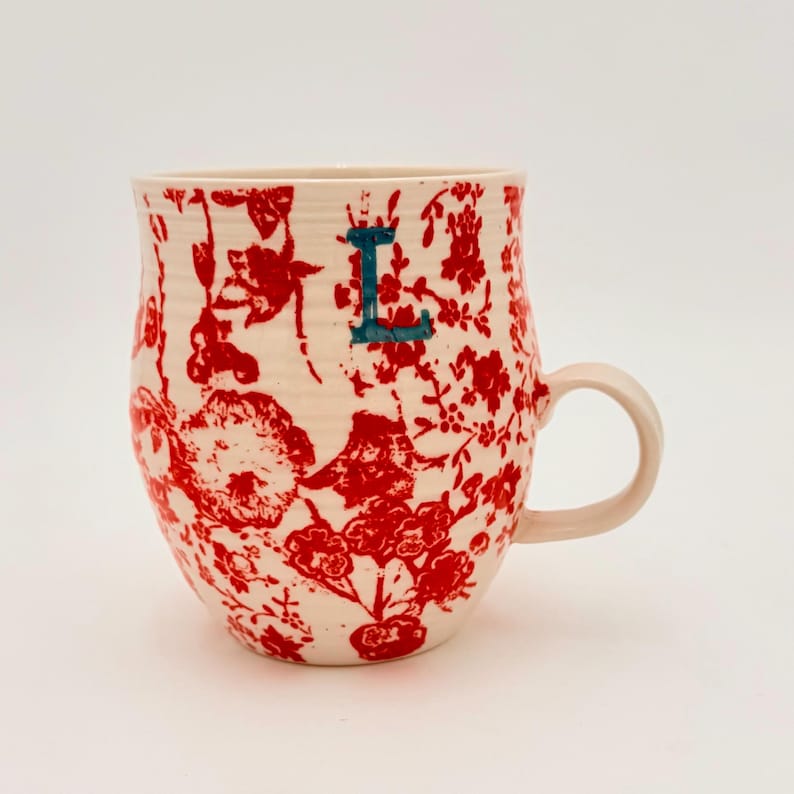 Może przedstawiać: Kremowy ceramiczny kubek z czerwonym kwiatowym wzorem. Kubek ma jasnor&oacute;żowy uchwyt i turkusową literę "L". Wz&oacute;r to powtarzający się motyw kwiat&oacute;w i liści. Unikalny element zastawy stołowej.