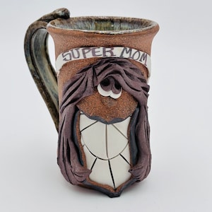 May include: A brown ceramic mug with a white and black face and the words "Super Mom" on a white banner. The mug has a brown handle and a brown glaze.