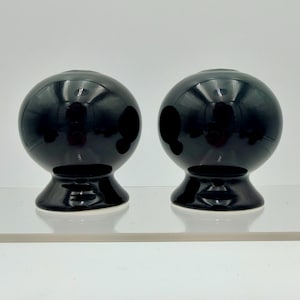 Puede incluir: Dos esferas decorativas de cerámica negra brillante. Cada esfera se asienta sobre una base acampanada. Las esferas reflejan la luz, creando reflejos y sombras. Los objetos se exhiben sobre una superficie transparente.
