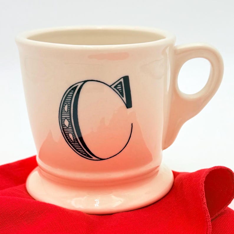 Puede incluir: Una taza de cer&aacute;mica color crema con una gran letra "C" negra en la parte delantera. La taza tiene una base redondeada, un asa y est&aacute; sobre una tela roja. La letra "C" est&aacute; estilizada con l&iacute;neas y formas decorativas.