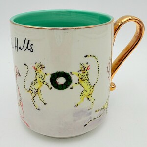 K&ouml;nnte beinhalten: Wei&szlig;er Keramikbecher mit goldenem Henkel und Rand. Der Innenraum ist hellgr&uuml;n. Der Becher zeigt Illustrationen von gelben Leoparden mit schwarzen Flecken, roten Schleifen und einem gr&uuml;nen Kranz. Das Wort "Halls" ist auf der Seite geschrieben.