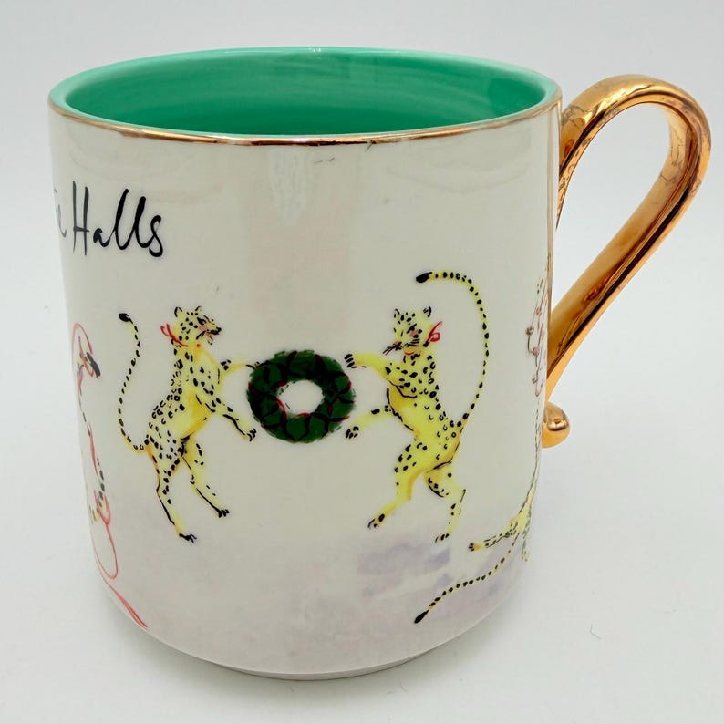 K&ouml;nnte beinhalten: Wei&szlig;er Keramikbecher mit goldenem Henkel und Rand. Der Innenraum ist hellgr&uuml;n. Der Becher zeigt Illustrationen von gelben Leoparden mit schwarzen Flecken, roten Schleifen und einem gr&uuml;nen Kranz. Das Wort "Halls" ist auf der Seite geschrieben.