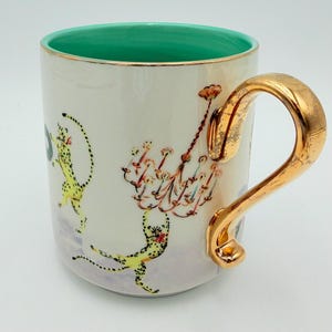 K&ouml;nnte beinhalten: Wei&szlig;e Keramiktasse mit goldenem Henkel und Rand, mit t&uuml;rkisfarbenem Innenraum. Die Tasse ist mit Illustrationen von gelben Geparden und einem Kronleuchter-Design verziert. Der Griff hat eine strukturierte, metallisch goldene Oberfl&auml;che.