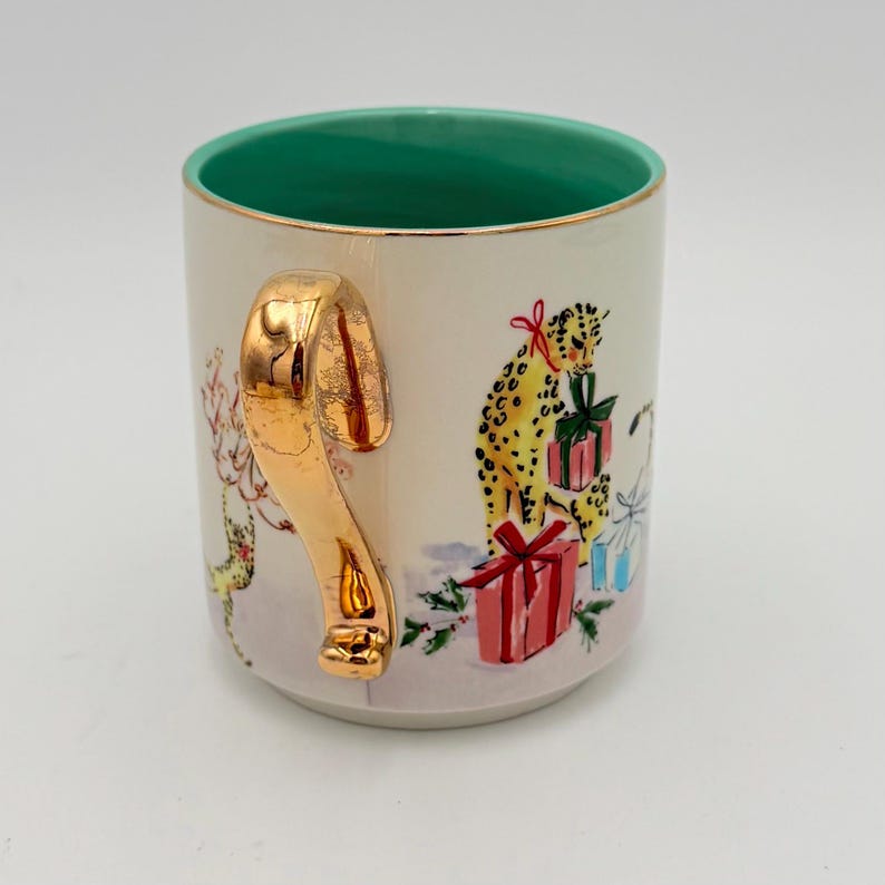K&ouml;nnte beinhalten: Cremefarbene Keramiktasse mit goldenem Henkel und Rand. Der Innenraum ist leuchtend gr&uuml;n. Die Tasse hat ein festliches Design mit einem Leoparden, verpackten Geschenken und einer Winterszene. Ein einzigartiges Weihnachtsgeschenk.