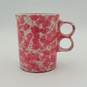 Può includere: Una tazza cilindrica in ceramica con base bianca e un vivace motivo a macchie di vernice rosa. La tazza si distingue per un design unico del manico, composto da due anelli circolari sovrapposti sul lato del recipiente.