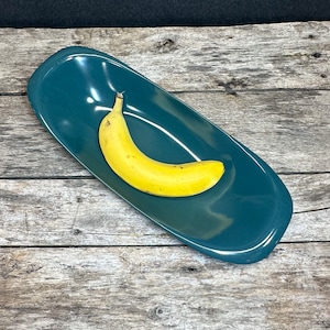 May include: A teal blue ceramic serving dish with a banana on top. The dish is oval shaped and has a glossy finish.