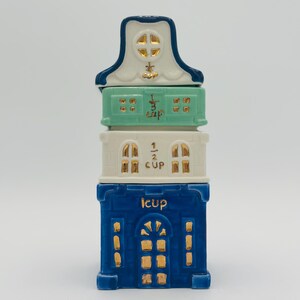 Vintage Anthropologie Sugar Cookie Townhouse Stacking Measuring Cups – Discontinued Holiday Kitchen Decor. A blue and white ceramic measuring cup set shaped like a house. The set includes four cups: 1 cup, 1/2 cup, 1/3 cup, and 1/4 cup.