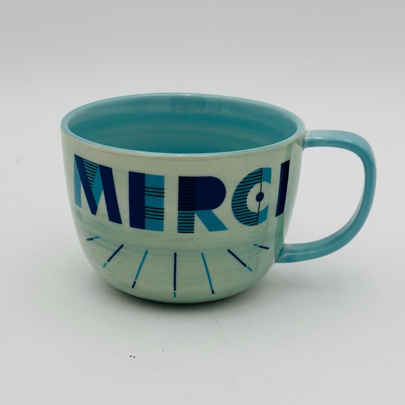 May include: A light blue ceramic mug with a white interior and a blue and white geometric design that says "MERCI" on the front.