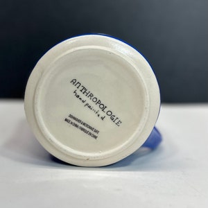 May include: A white ceramic mug with a blue exterior and a white interior. The bottom of the mug features the text "ANTHROPOLOGIE hand painted" and "DISHWASHER & MICROWAVE SAFE MADE IN CHINA / FABRIQU&Eacute; EN CHINE."