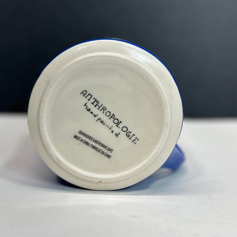 May include: A white ceramic mug with a blue exterior and a white interior. The bottom of the mug features the text "ANTHROPOLOGIE hand painted" and "DISHWASHER & MICROWAVE SAFE MADE IN CHINA / FABRIQU&Eacute; EN CHINE."