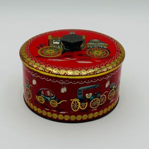 May include: A round red metal tin with a removable lid featuring a black hexagonal knob. The exterior is decorated with gold scrollwork borders and illustrations of horse-drawn carriages in blue, yellow, and black colors around the sides.