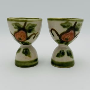 Könnte beinhalten: Zwei Eierbecher aus Keramik mit beigem Sockel und grünem Rand. Jeder Becher hat ein handgemaltes Design einer rötlich-orangen Birne mit grünen Blättern und braunen Stielen. Die Eierbecher haben eine einzigartige Sanduhrform.