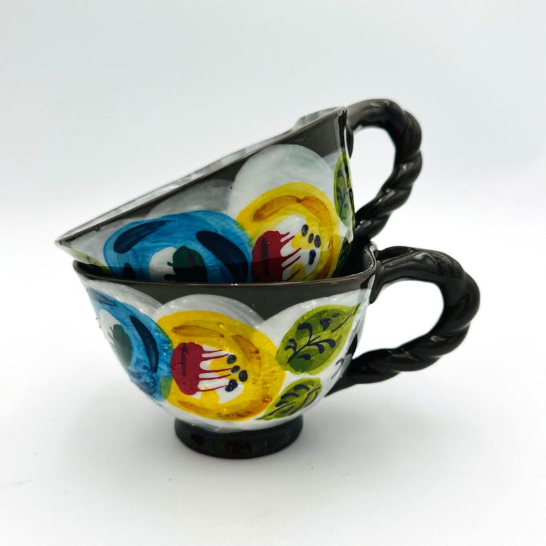 Puede incluir: Dos tazas de t&eacute; de cer&aacute;mica apiladas con dise&ntilde;os florales en amarillo, azul y rojo. Las tazas tienen asas y bordes negros trenzados. El patr&oacute;n floral incluye hojas verdes y detalles blancos.