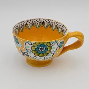 May include: A yellow ceramic teacup with a floral design. The cup features a large flower in shades of blue and green, with a white background. The rim is decorated with a scalloped pattern. The handle and base are also yellow.