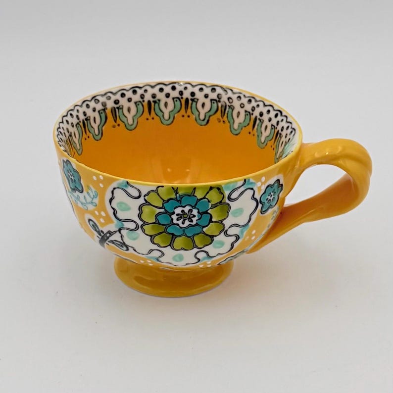 May include: A yellow ceramic teacup with a floral design. The cup features a large flower in shades of blue and green, with a white background. The rim is decorated with a scalloped pattern. The handle and base are also yellow.