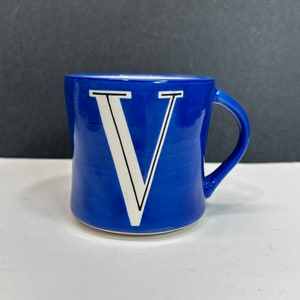 May include: A cobalt blue ceramic mug with a white letter "V" in a bold, serif font. The mug has a curved handle and a slightly irregular shape, giving it a handmade appearance. The background is a neutral gray and white.