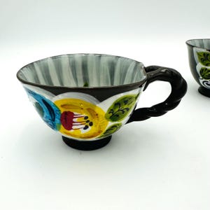 Puede incluir: Un par de tazas de t&eacute; de cer&aacute;mica con dise&ntilde;os florales. Las tazas tienen un borde y un asa negros, con un colorido estampado floral en amarillo, azul y rojo. El interior de la taza tiene rayas verticales grises y blancas.