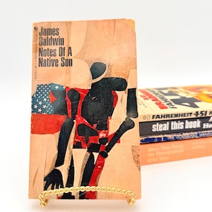 Puede incluir: Libro de bolsillo vintage titulado "Notes Of A Native Son" de James Baldwin. La portada presenta una ilustración estilo collage con una figura negra y roja, y una bandera estadounidense. Otros libros están apilados en el fondo.