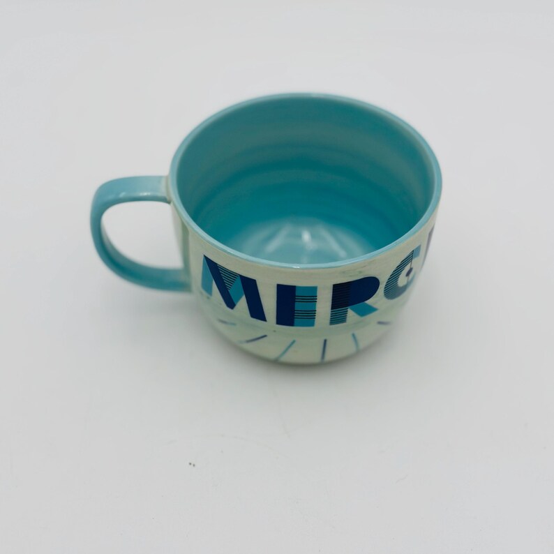 May include: A light blue ceramic mug with a white interior and a graphic of the word "MERCI" in blue and white.