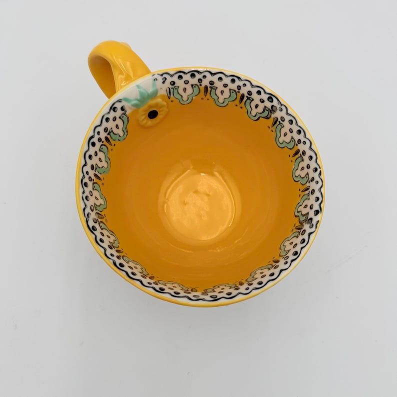 May include: Overhead view of a yellow ceramic teacup with a handle. The cup's interior is a solid yellow, while the rim features a decorative border of white, black, and green floral designs. A small floral accent is on the cup's exterior.