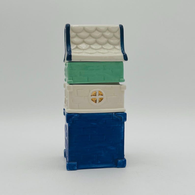 May include: A stack of four ceramic houses in blue, white, and green. The top house has a white roof with a blue trim and a scalloped design. The second house is green, the third is white with a gold window, and the bottom house is blue.