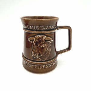 May include: A brown glazed ceramic mug featuring a raised relief of a bull head on the front. The text THE MUSTARD SHOP is embossed near the top rim, and the text NORWICH-ENGLAND is embossed near the base. The mug has a single side handle.