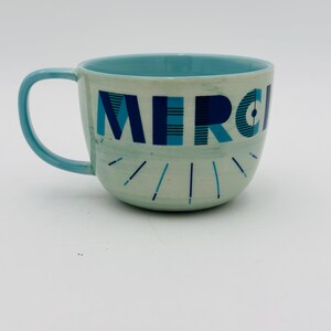 May include: A light blue ceramic mug with a white interior and a blue and white geometric design that says "MERCI" on the front.