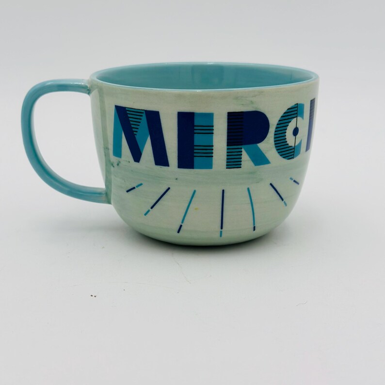 May include: A light blue ceramic mug with a white interior and a blue and white geometric design that says "MERCI" on the front.