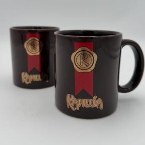 May include: Two dark brown ceramic mugs with the Kahlúa logo. Each mug features a gold-colored seal with a letter "K" on a red banner. The Kahlúa name is printed in gold script below the banner.