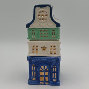 May include: A three-tiered ceramic house figurine with a blue, white, and green color scheme. The bottom tier is blue with gold accents and the text "BOULANGERIE". The middle tier is white with a gold star and gold windows. The top tier is green with gold windows.