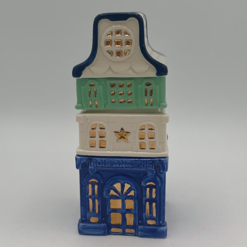 May include: A three-tiered ceramic house figurine with a blue, white, and green color scheme. The bottom tier is blue with gold accents and the text "BOULANGERIE". The middle tier is white with a gold star and gold windows. The top tier is green with gold windows.